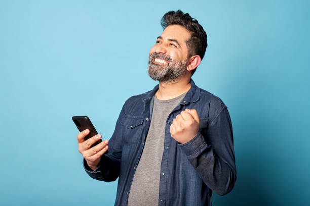 Mid latin adult man smiling celebrating good news with fist raised, holding mobile phone. Indoor studio shot isolated on blue background. Copy space.