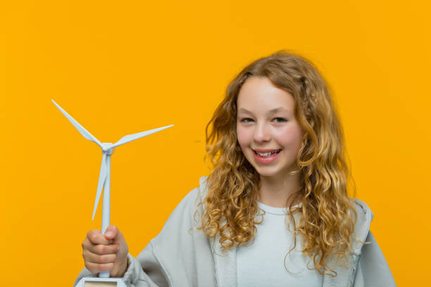 Happy teenage girl wearing grey hoodie and white top holding wind turbine model and smiling at camera. Studio shot, yellow background.